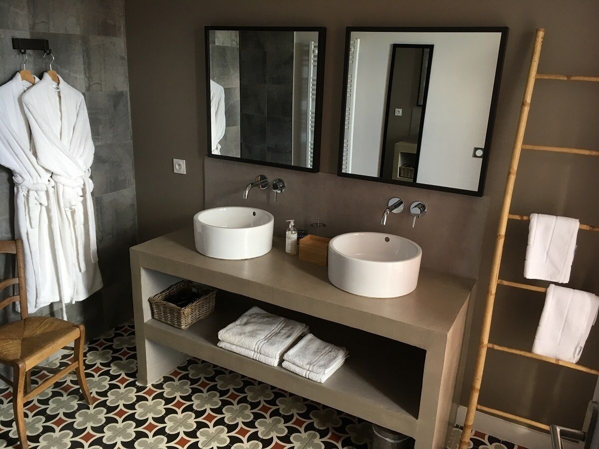 A bathroom with two round sinks, a mirror, a wooden chair, and a shelf with towels.