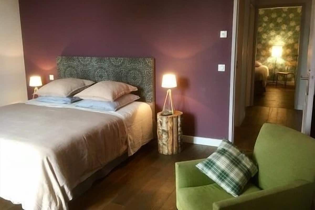 A hotel room with a bed, two bedside lamps, a green armchair, and a view into the adjacent room.