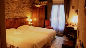 A cozy room with a brick wall, wooden ceiling beams, a double bed with a white bedspread, a wooden wardrobe, a chair, and a small table with a lamp. - La Maréchalerie (Monthodon)