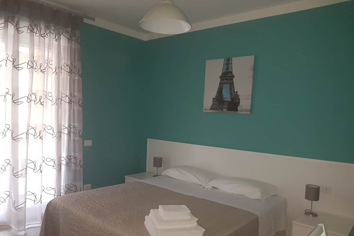 A bedroom with a bed, a nightstand, a lamp, a painting of the Eiffel Tower, and a window with sheer curtains.