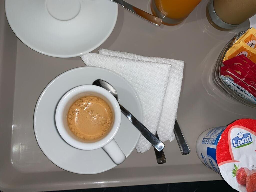 Ein Frühstücksbrett mit einer Tasse Kaffee, einem Teller, einem Servietttuch, einem Löffel, einem Glas Orangensaft und einem Joghurtbecher.