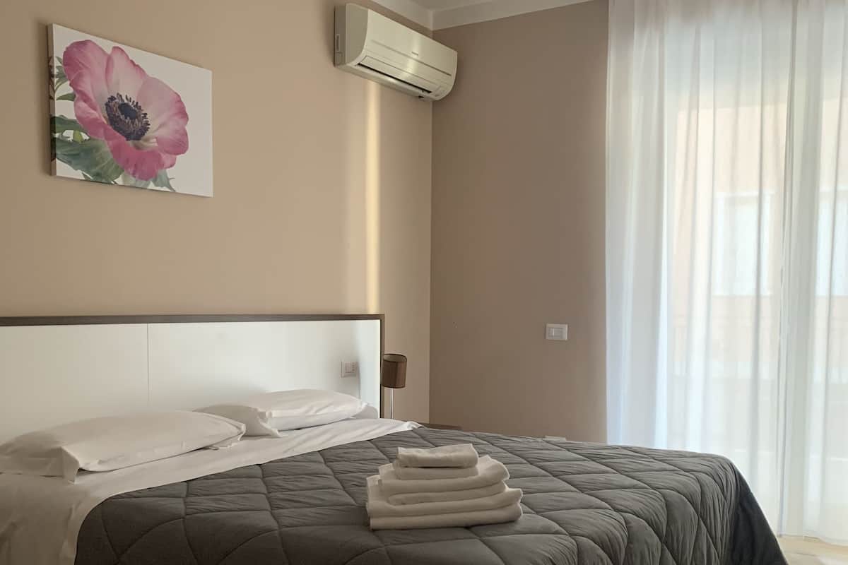 A neatly made bed with a grey quilt, a white pillow, and a folded towel on top. There is a wall-mounted air conditioner and a painting of a flower above the bed.