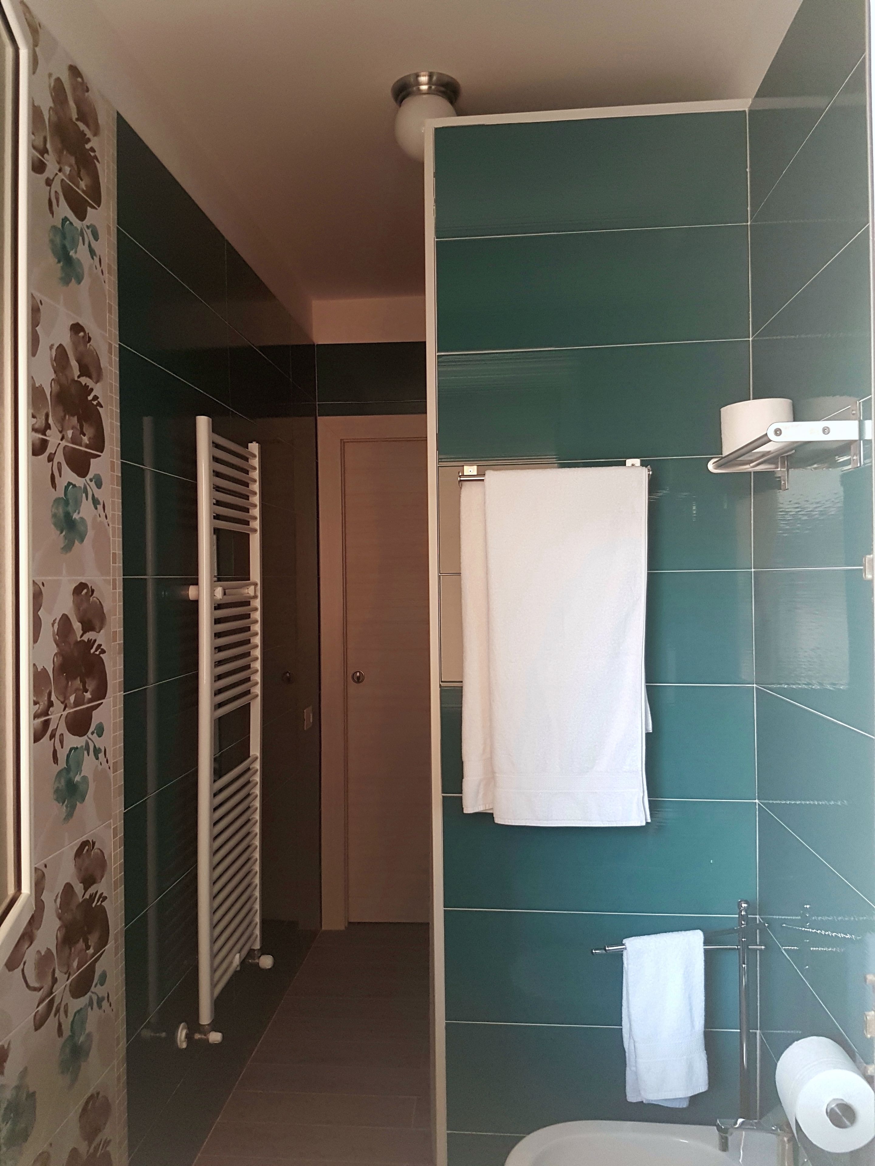 Un baño con azulejos turquesa, una toalla blanca colgada en un perchero, un porta papel higiénico y una zona de ducha.