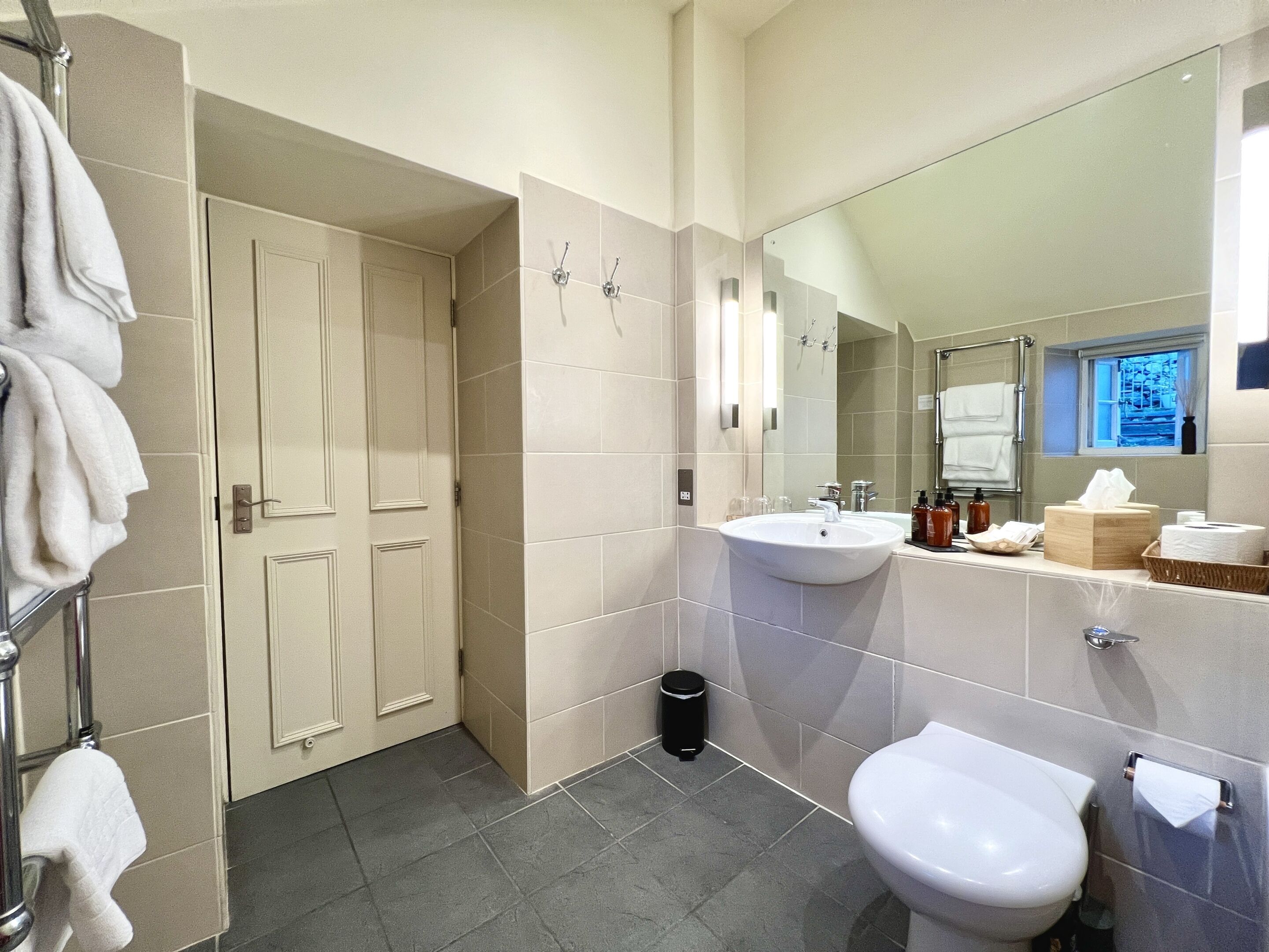 A modern bathroom with a white sink, a toilet, and a mirror. There is a towel rack with towels and a trash can.