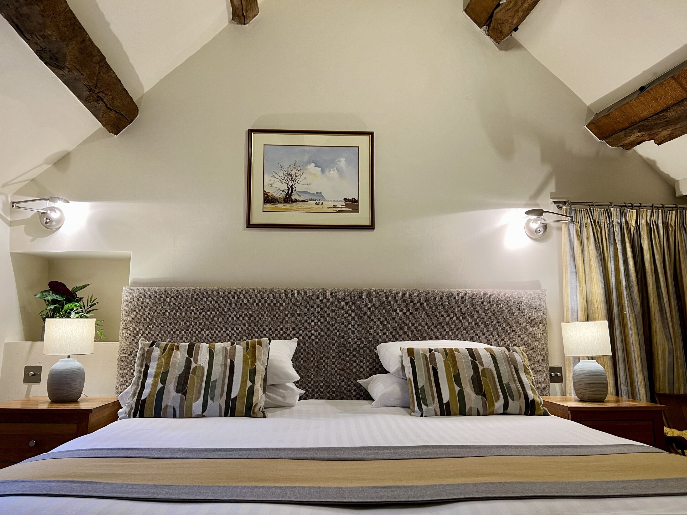 A bedroom with a large bed, two nightstands, a painting on the wall, and a window with curtains.