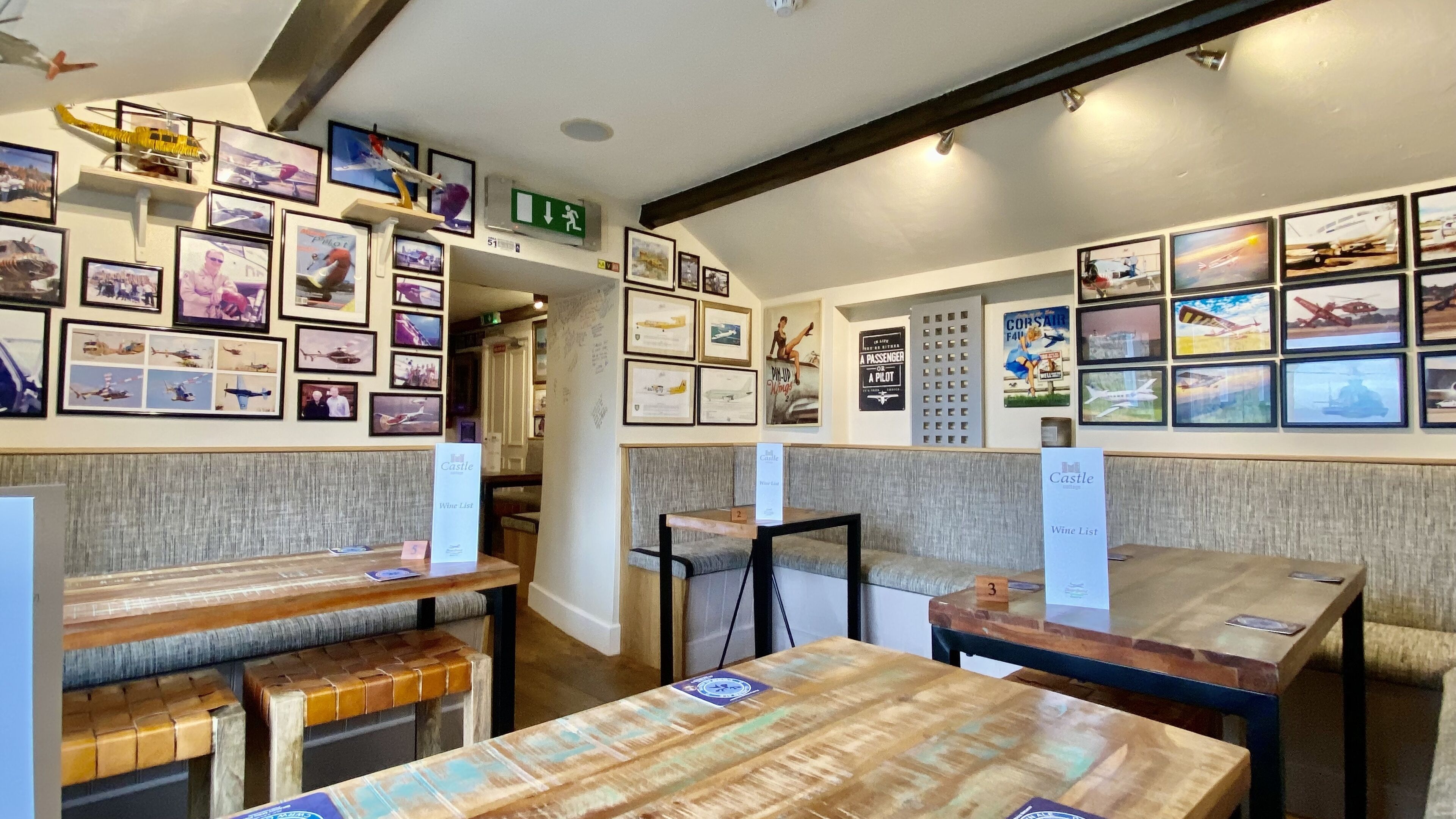 Un restaurant avec des tables et des bancs en bois, des tableaux sur le thème de l’aviation accrochés aux murs et un panneau de sortie vert.