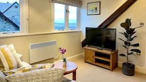 A cozy living room with a large TV, a comfortable sofa, a small table with a plant, and a view of the outside through a window. - Castle Cottage Inn (Harlech)