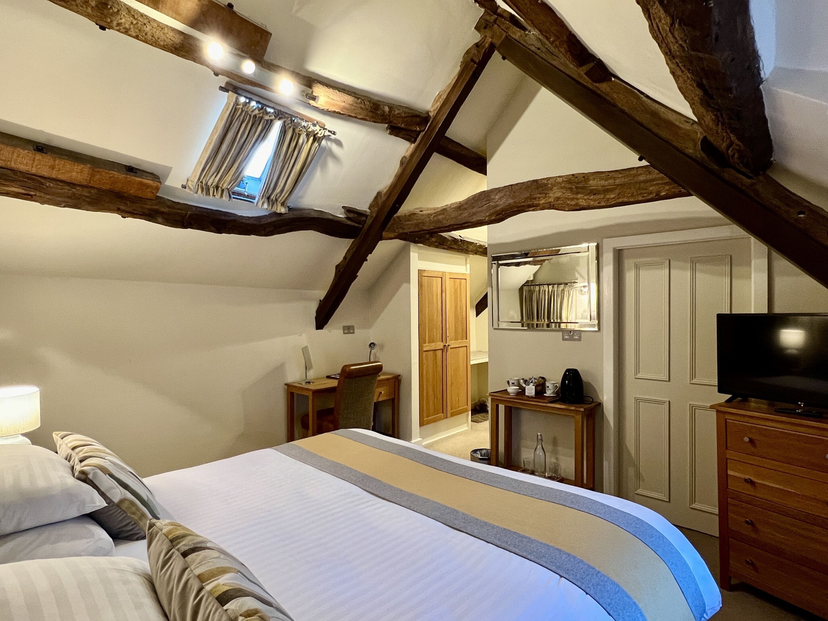 A bedroom with a large bed, wooden beams, a desk, a mirror, and a television.