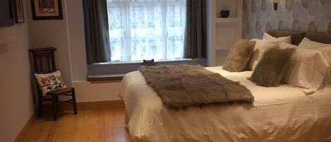 A bedroom with a large bed, a window with curtains, a chair, and a small table.