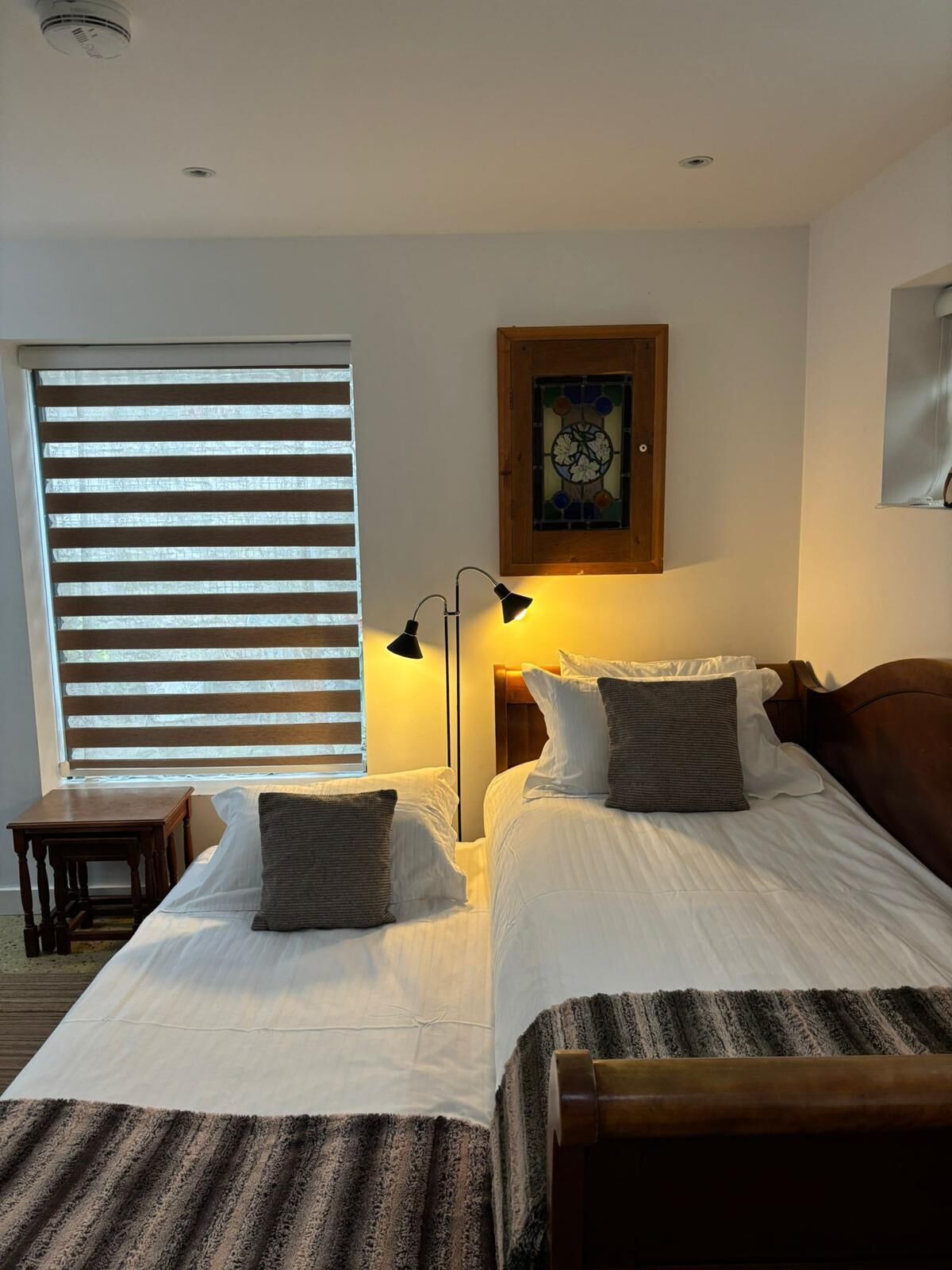 A hotel room with two beds, a nightstand, a lamp, a window with blinds, and a framed artwork on the wall.