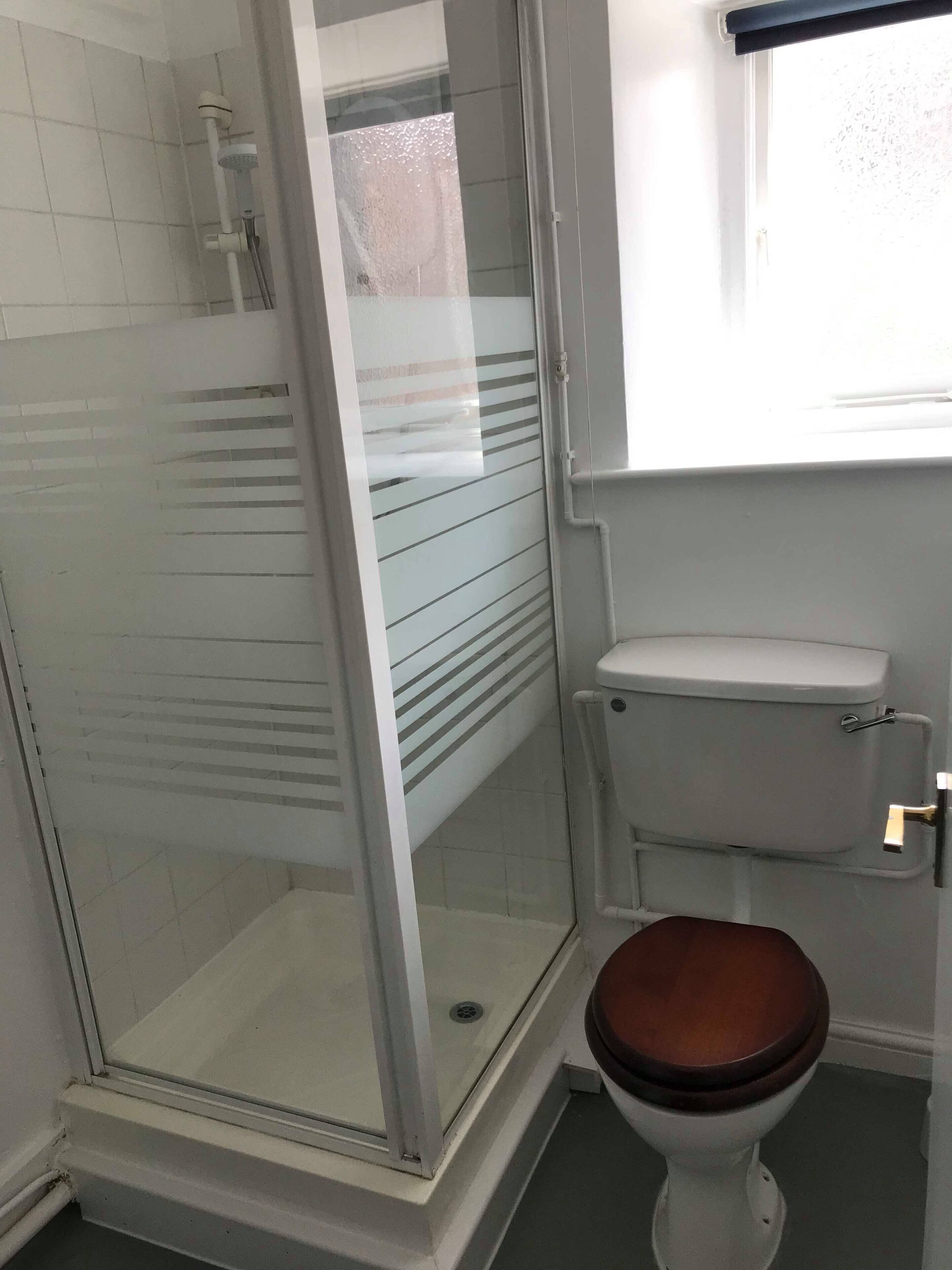 A bathroom with a glass-enclosed shower, a toilet with a wooden seat, and a window above the toilet.