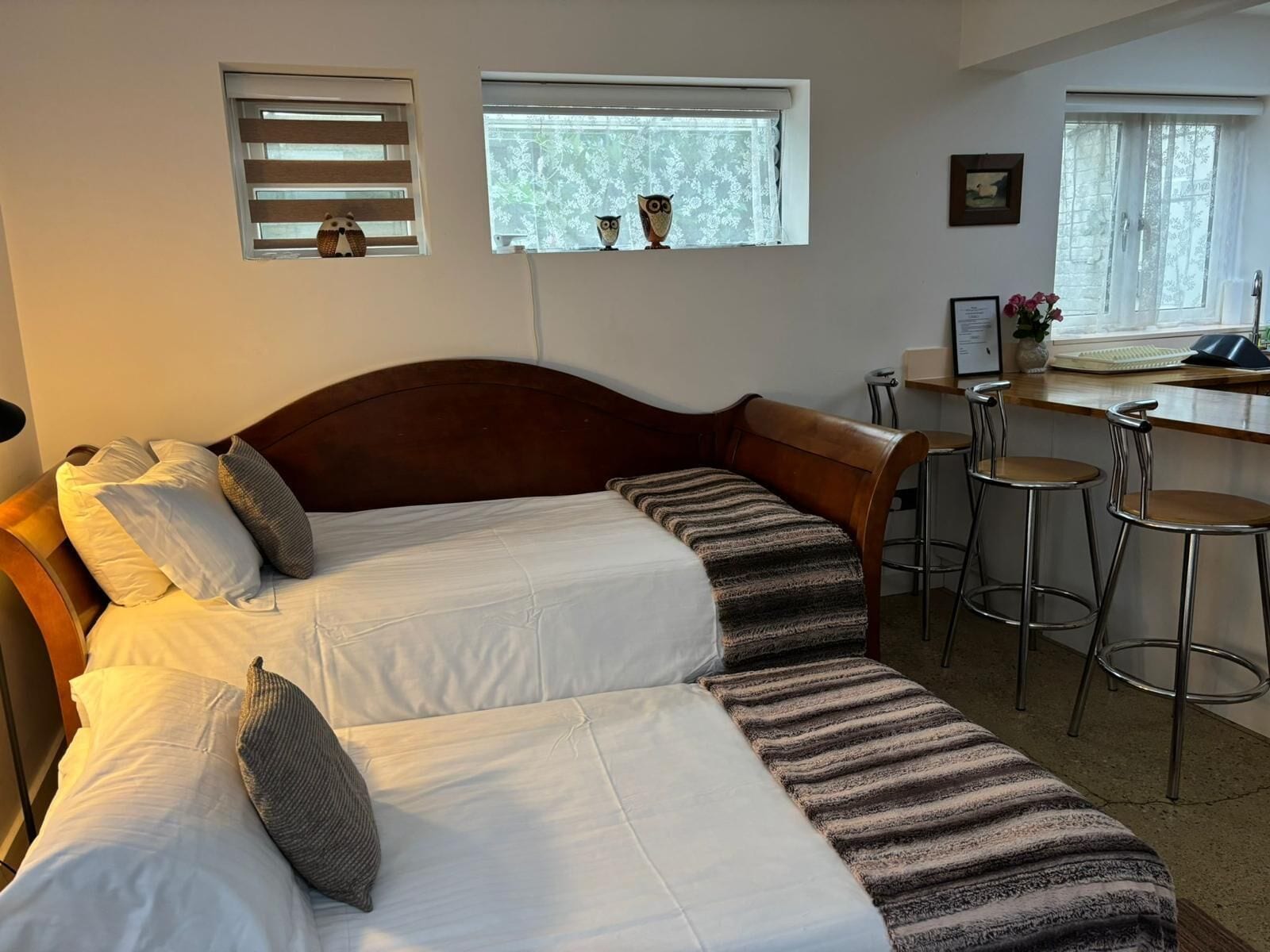 A bedroom with a bed, two windows with blinds, a desk with a chair, and a small bar stool.