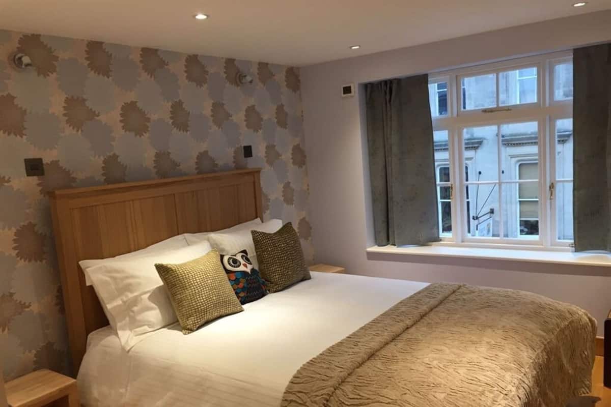 A neatly made bed with a wooden headboard, a window with curtains, and patterned wallpaper.