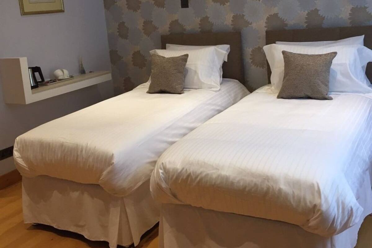 Two single beds with white linens and gray pillows, a small shelf with decorative items, and a patterned wallpaper on the wall.