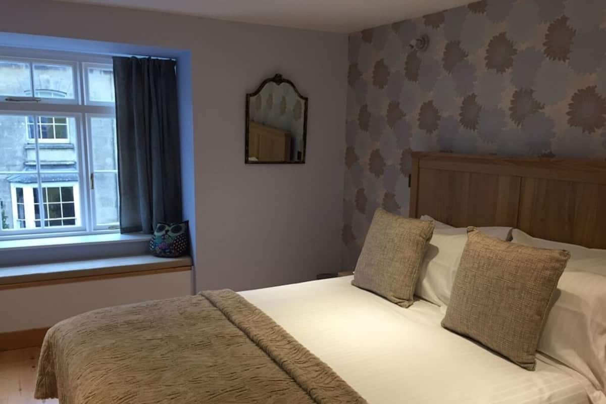 A bedroom with a large bed, a window with curtains, a mirror, and patterned wallpaper.