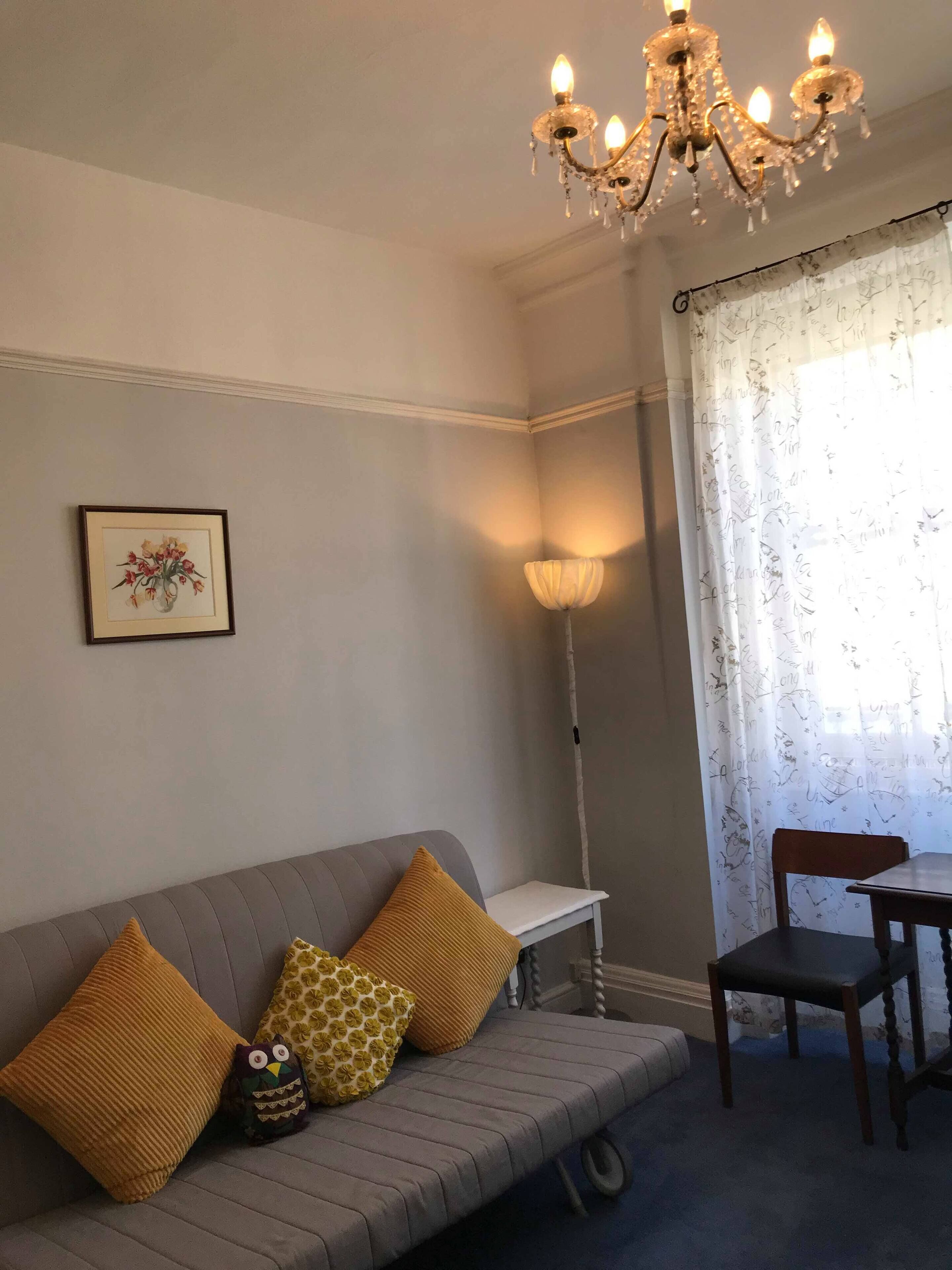 Una habitación con un sofá gris, una silla de madera, una araña, un cuadro enmarco y una lámpara.