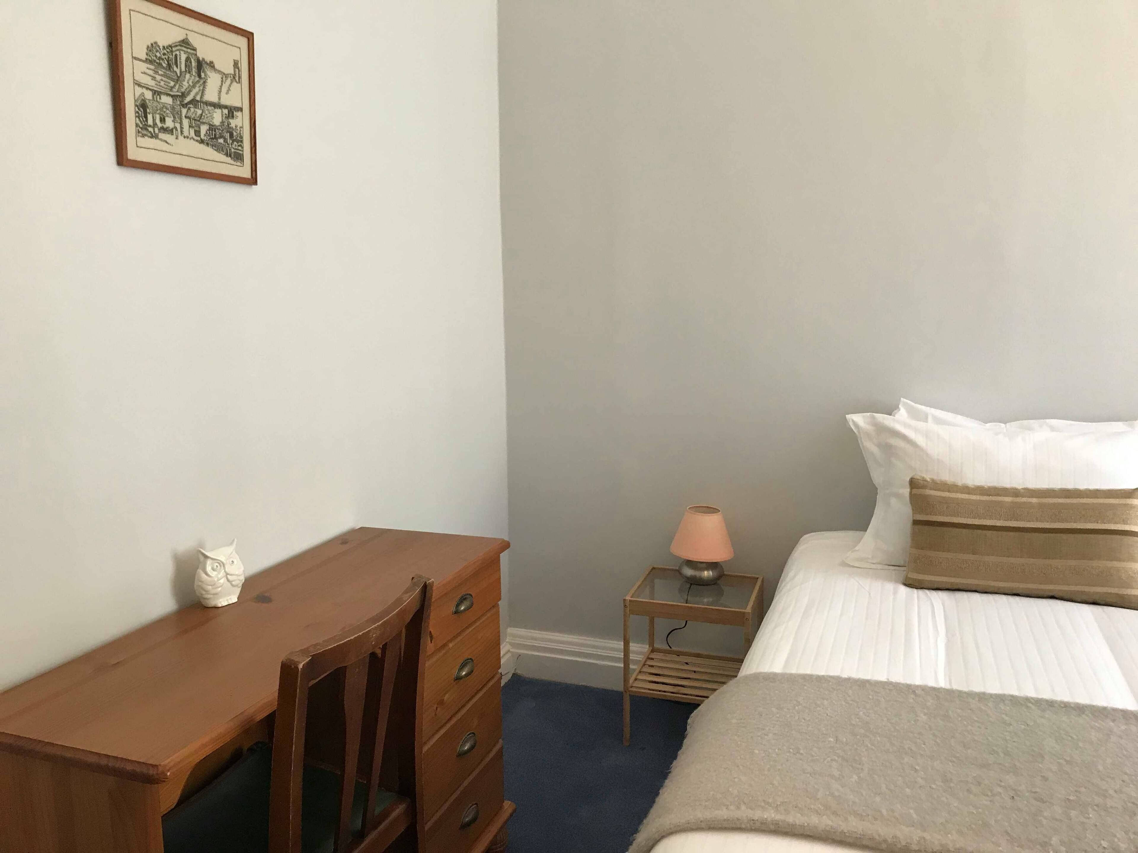 A bedroom with a bed, a wooden dresser, a nightstand with a lamp, and a framed picture on the wall.