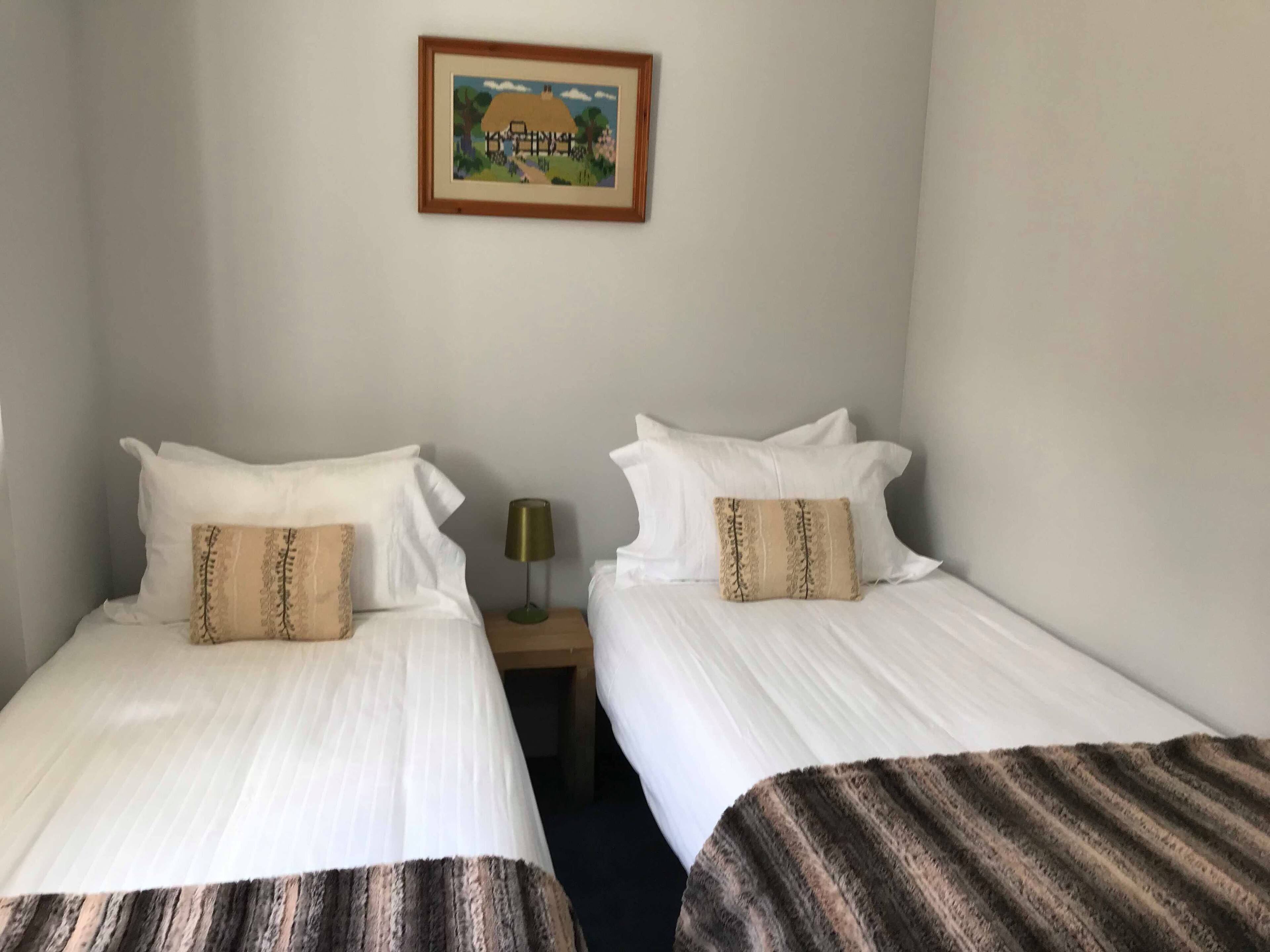 Ein Hotelzimmer mit zwei Betten, jeweils mit weißer Bettwäsche und einer gestreiften Decke. Dazwischen befindet sich ein Nachttisch mit einer Lampe, darüber an der Wand ein gerahmtes Bild.
