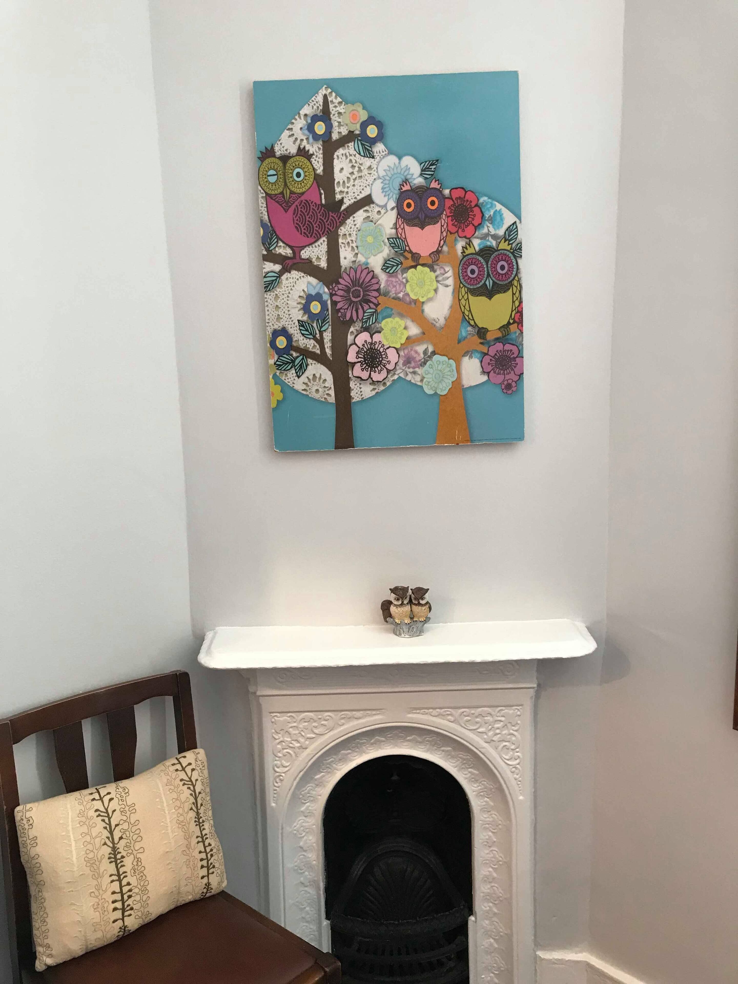 A room with a white fireplace, a white mantel, and a colorful painting of owls and flowers above it.