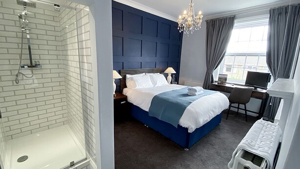 Una habitación de hotel con ducha envuelta en vidrio, una cama con cabecera azul, un escritorio con ordenador y una ventana con cortinas.
