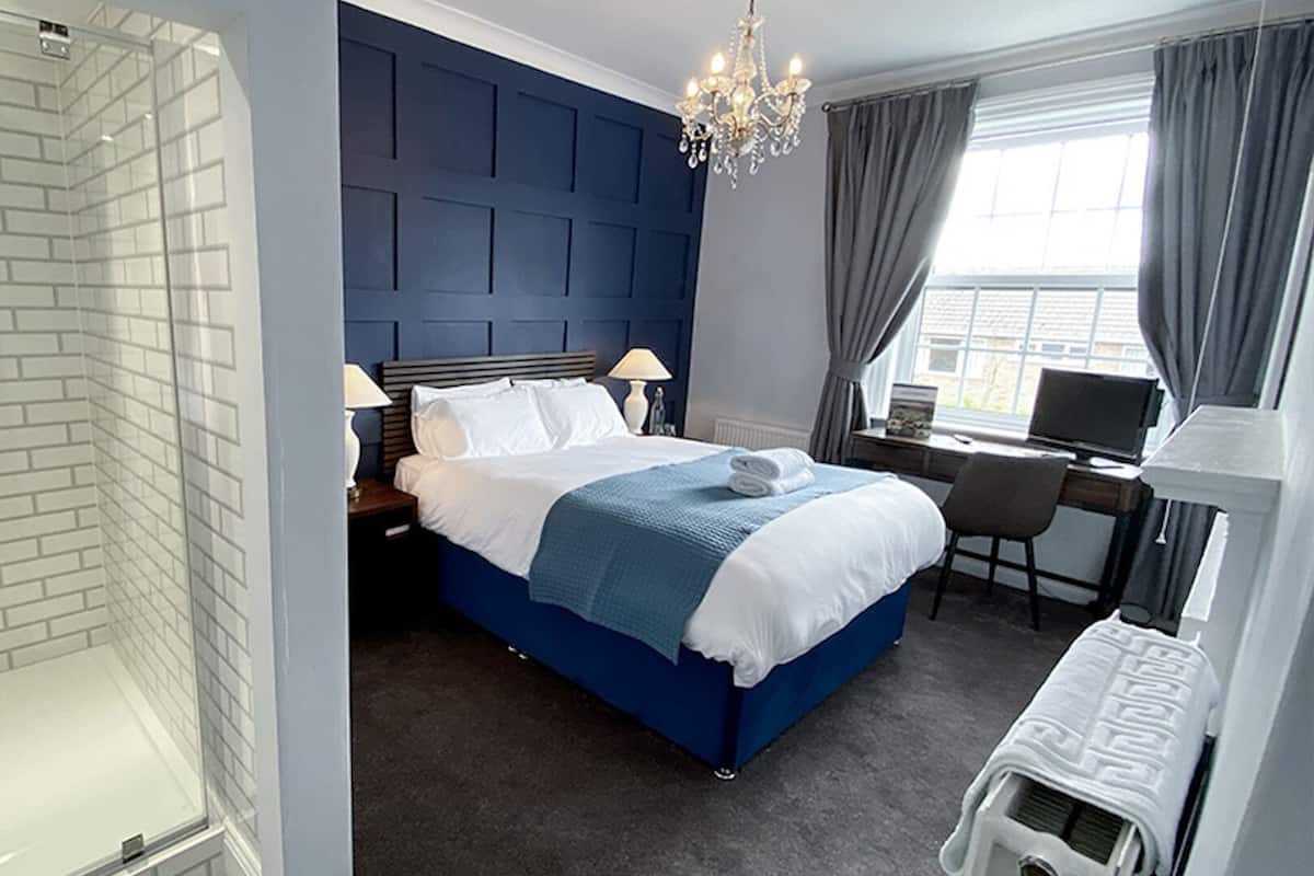 A hotel room with a glass-enclosed shower, a bed with a blue headboard, a desk with a computer, and a window with curtains.