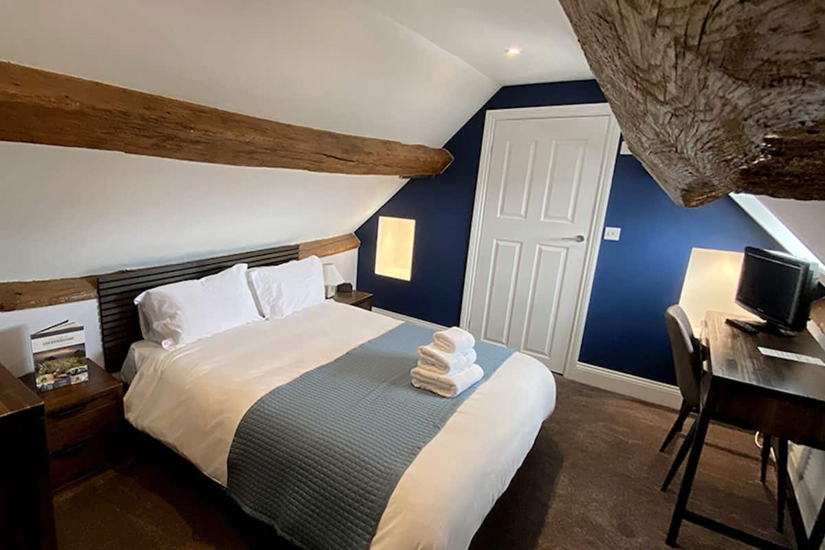 A hotel room with a bed, bedside table, lamp, and a desk with a computer.