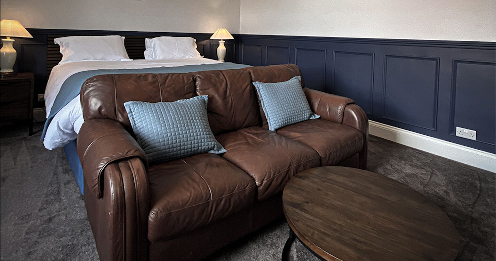 A brown leather sofa with two pillows, a matching ottoman, and a bed with white linens in a room with dark blue walls and a gray carpet.