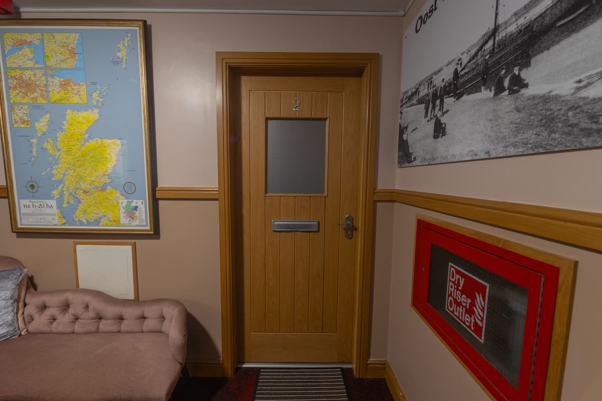 A hotel room with a wooden door, a framed map, a framed photograph, and a sign indicating a dry riser outlet.