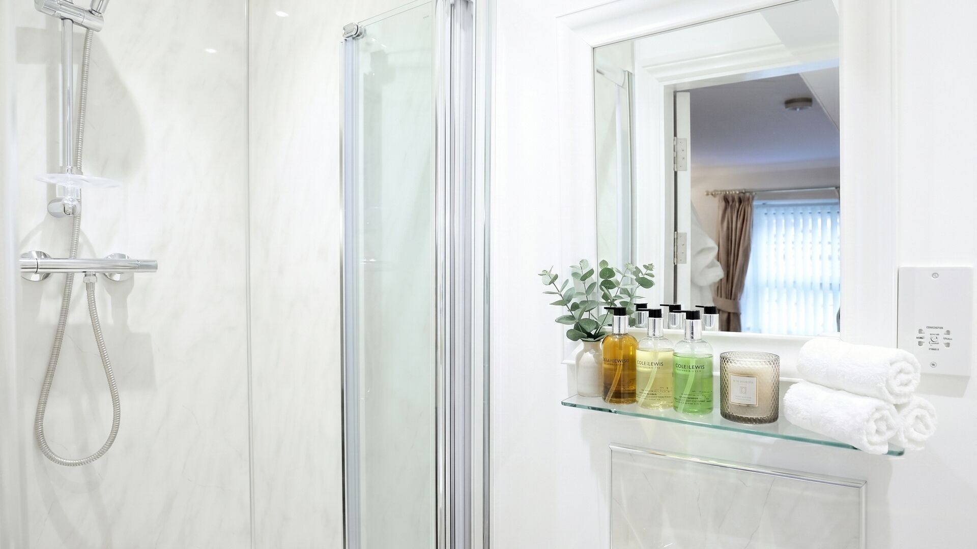A modern bathroom with a glass shower enclosure, a glass shelf with toiletries, and a mirror reflecting a room with a window and curtains.