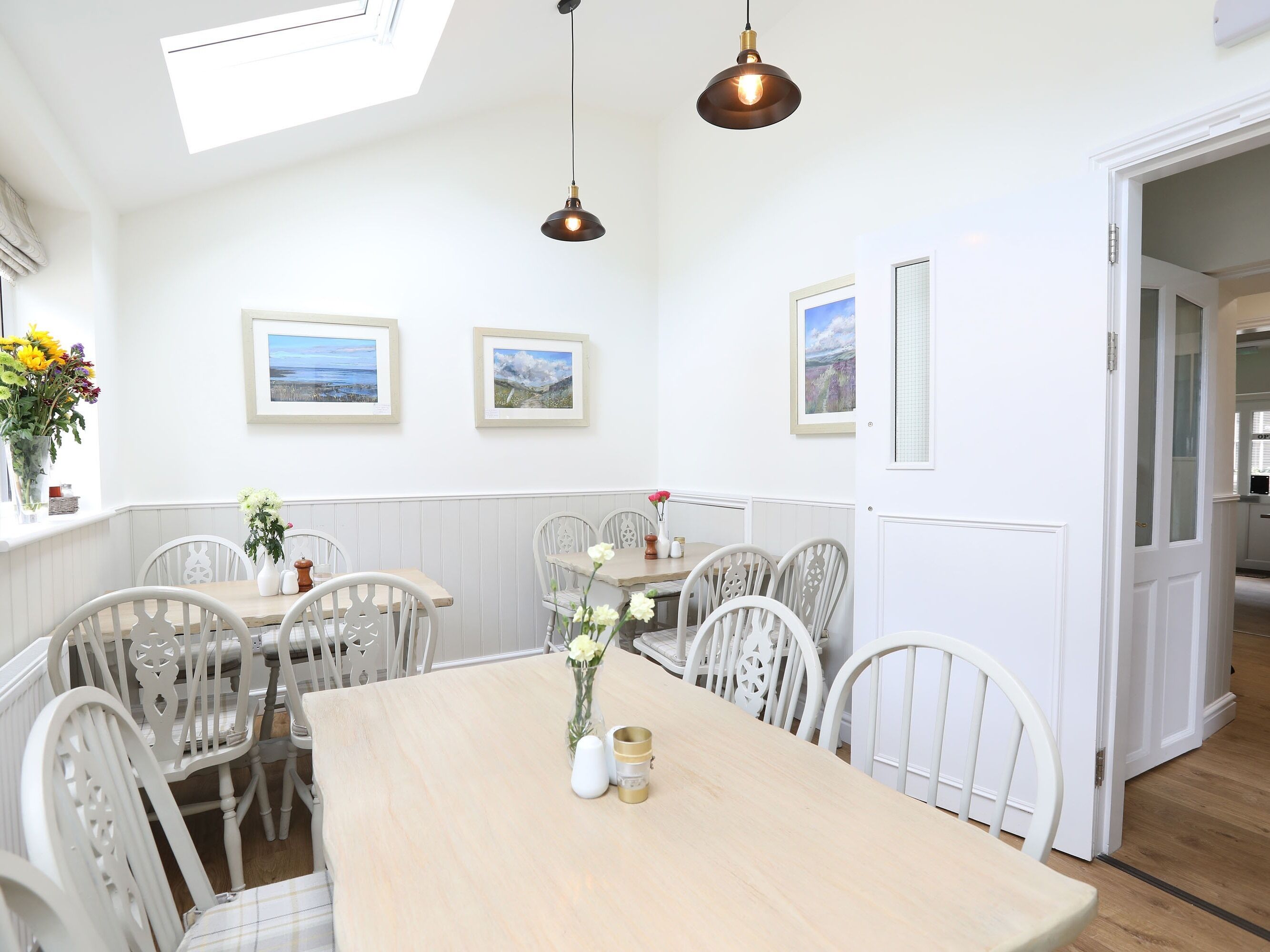 A dining area with a wooden table, white chairs, and pendant lighting. There are framed pictures on the wall and a window with a view.