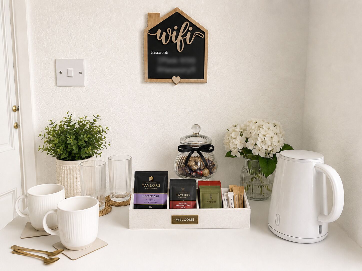 A neatly arranged coffee station with mugs, tea bags, and a kettle, a potted plant, and a wall-mounted sign with a wifi password.