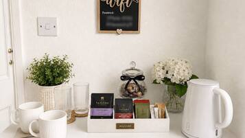 A neatly arranged coffee station with mugs, tea bags, and a kettle, a potted plant, and a wall-mounted sign with a wifi password.
