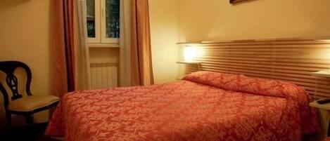 A room with a bed, a chair, a window with curtains, a wall-mounted picture, and a bedside lamp.