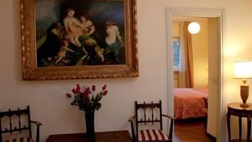A room with a painting, a vase of flowers, two chairs, a table, and a bed in the background.