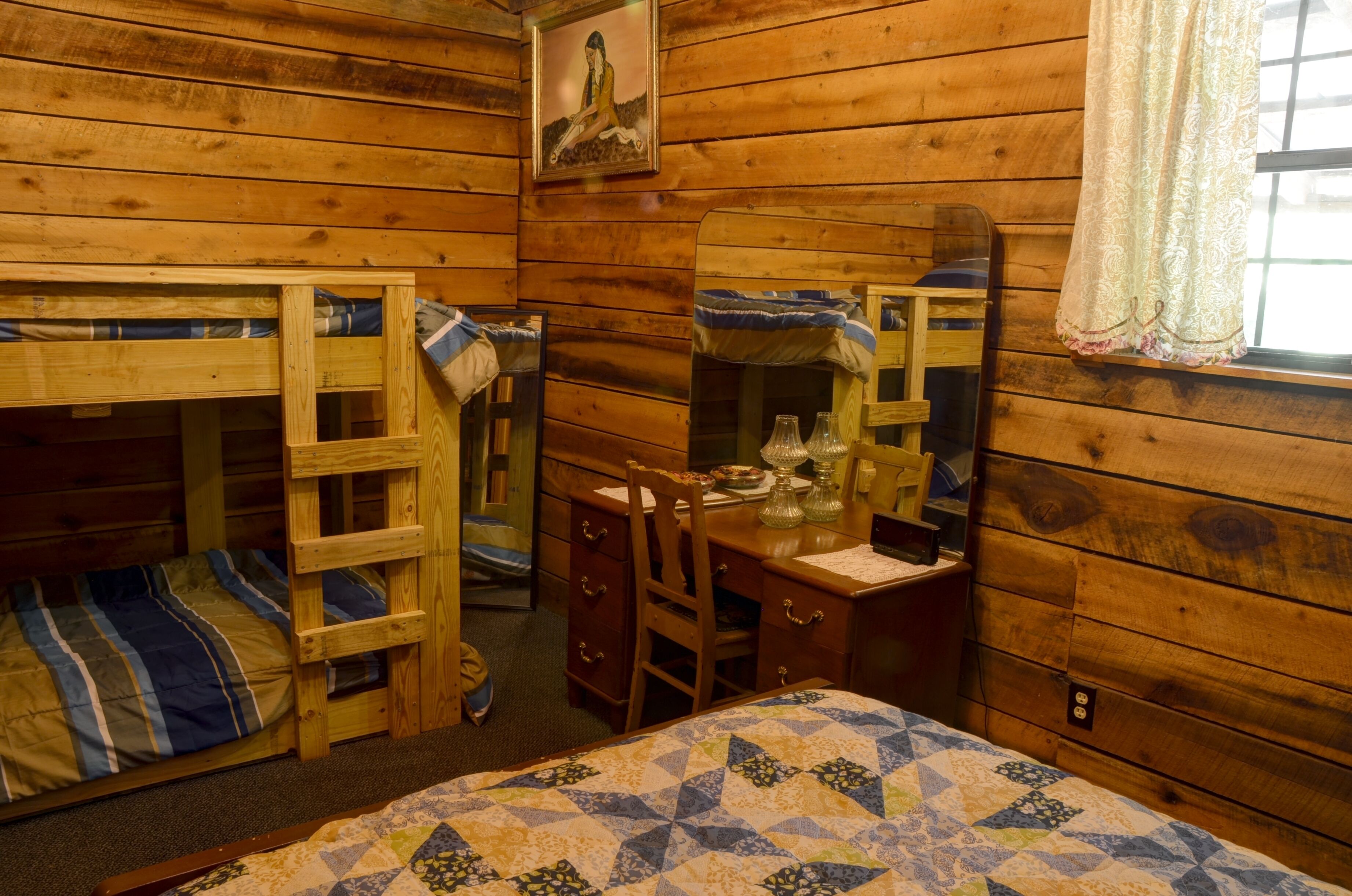 A cabin-style room with wooden walls, a bunk bed, a desk with a mirror, and a window with a curtain.