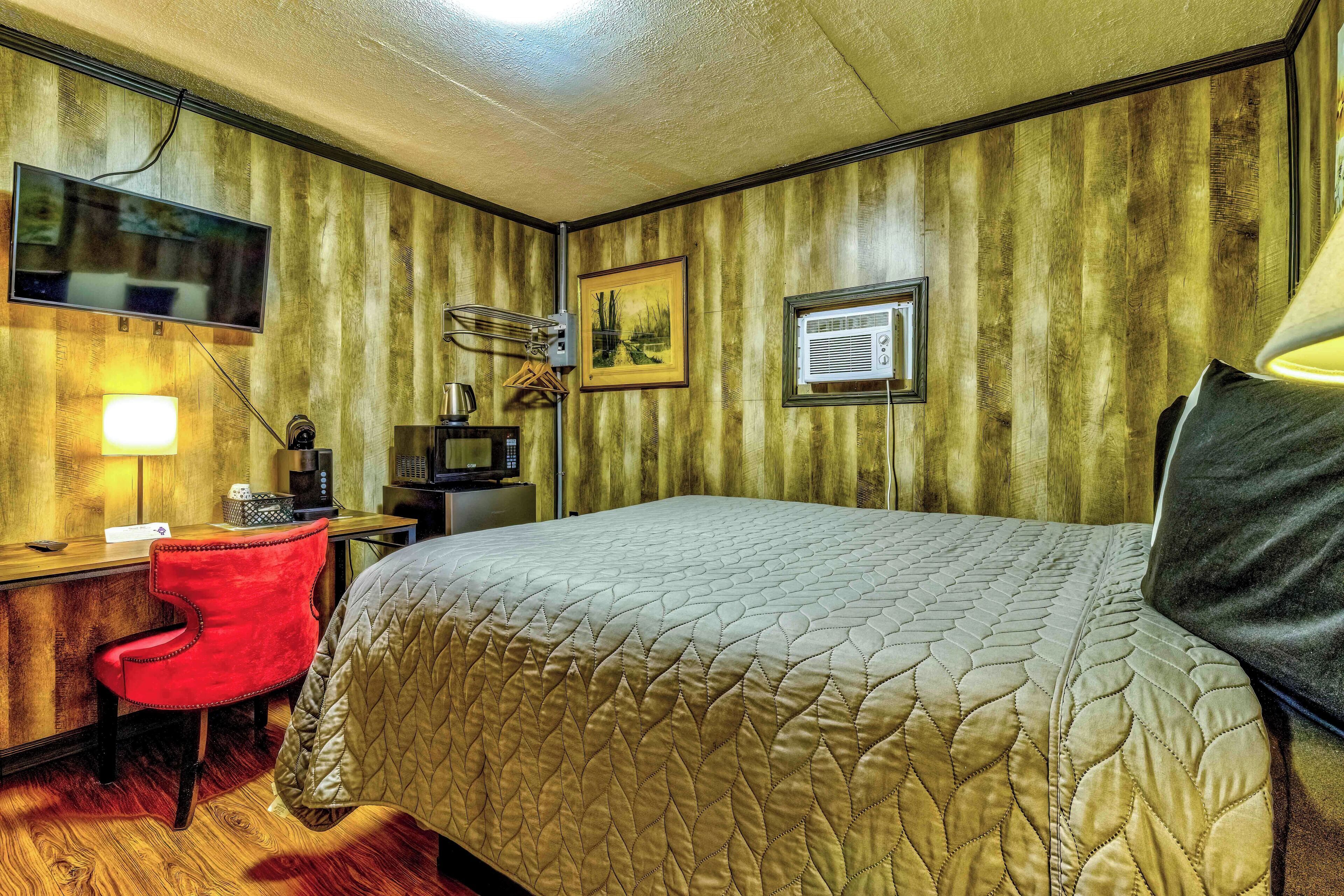 A hotel room with a bed, a desk with a chair, a television, a painting, and an air conditioning unit.