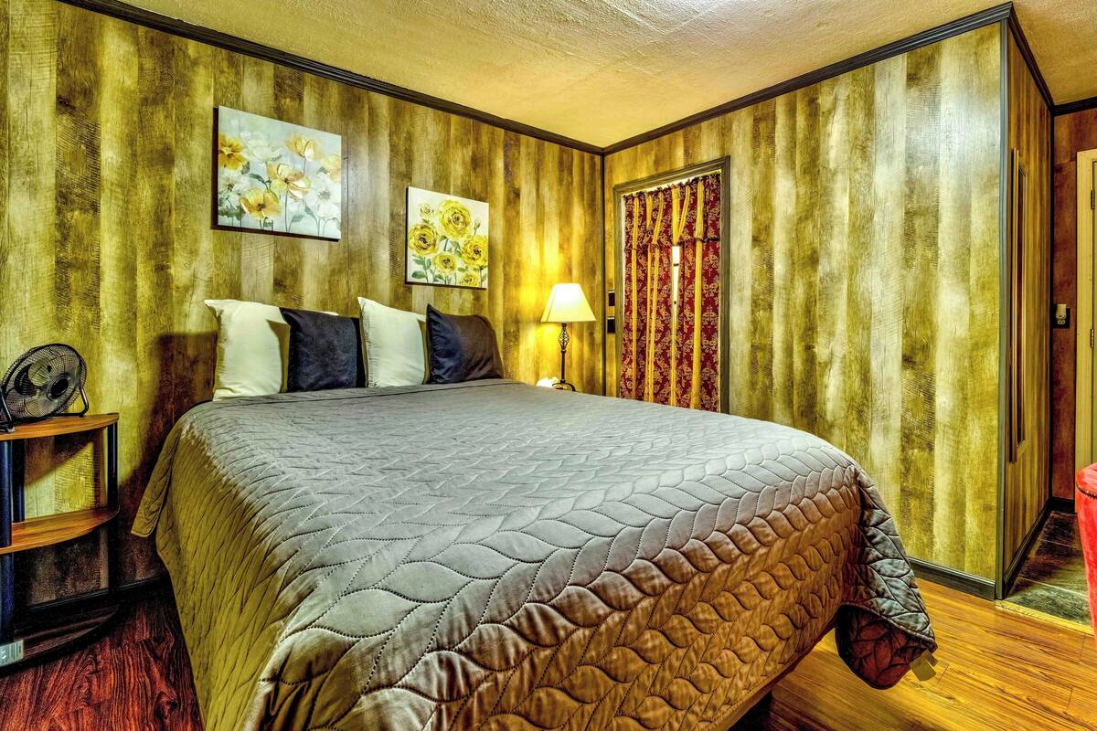 Una habitación con una cama grande, paneles de madera, dos cuadros, una cortina roja y una mesita de noche con lámpara.
