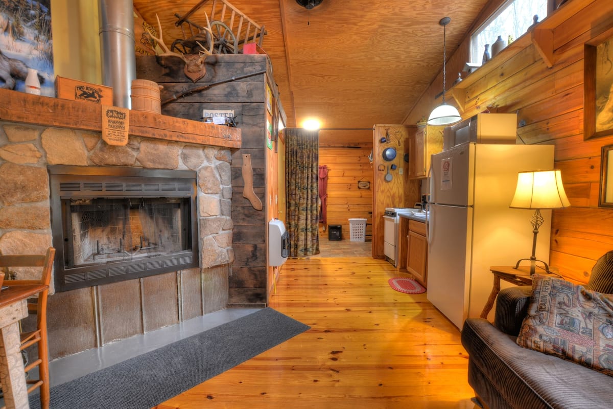 Un acogedor interior de cabaña con chimenea de piedra, pisos de madera, zona de comedor y cocina con refrigerador.