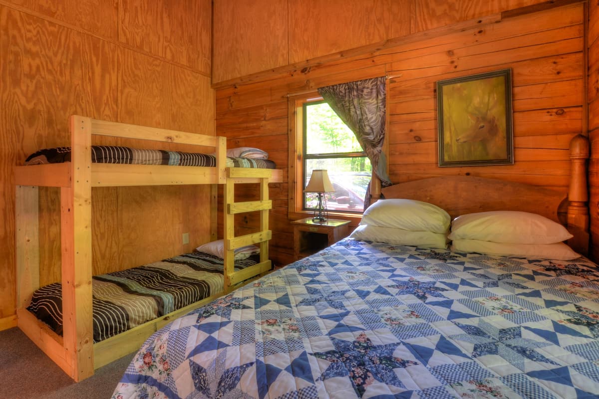 Habitación de cabaña de madera con literas, una cama con colcha, una mesita de noche, una lámpara y una ventana con cortinas.