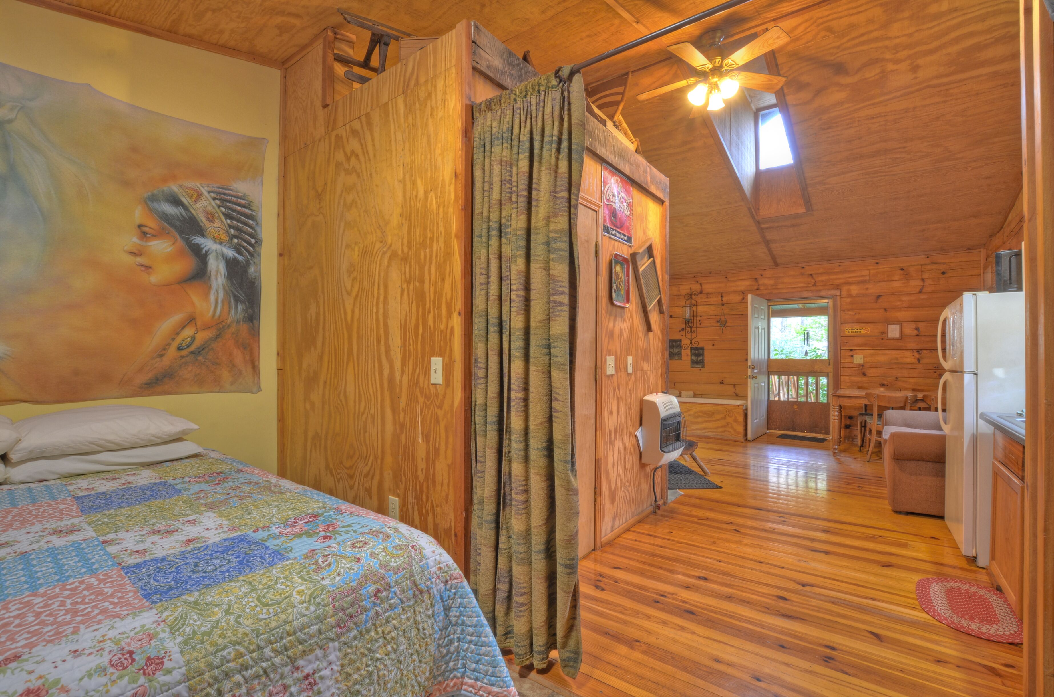 A cozy log cabin bedroom with a quilted bed, wooden walls, a ceiling fan, and a view to the dining area.
