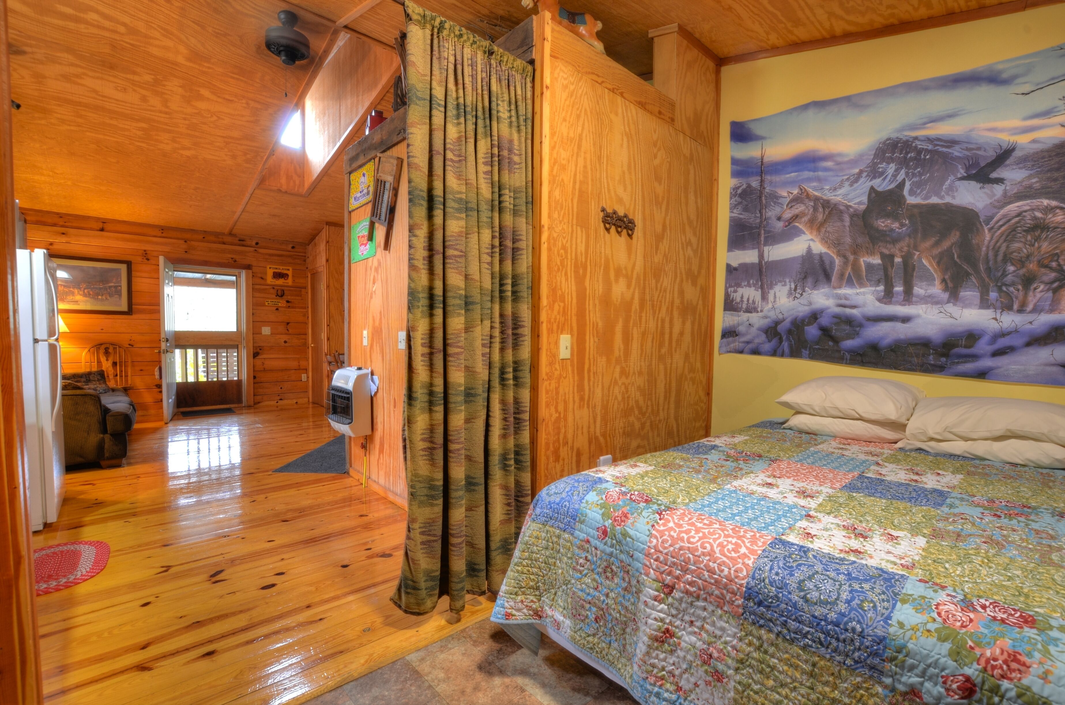 A cozy cabin room with a wooden ceiling, a quilted bed, a painting of wolves, and a wooden door.