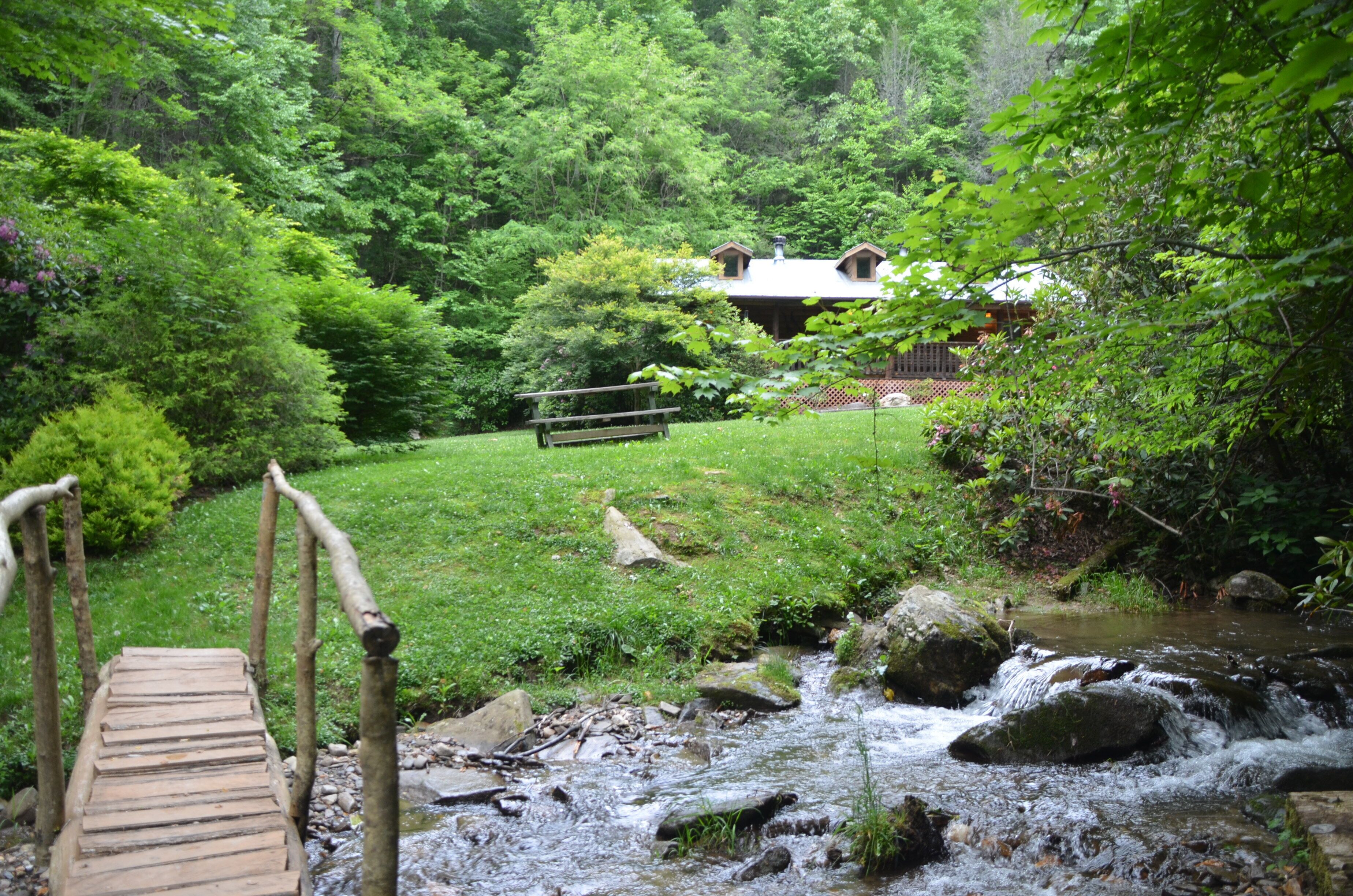 A wooden bridge over a stream leading to a house surrounded by lush greenery.