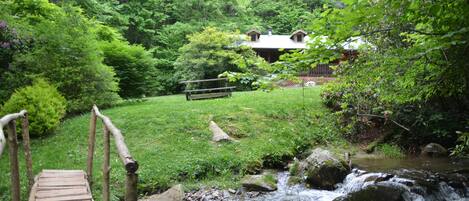 A wooden bridge over a stream leading to a house surrounded by lush greenery.