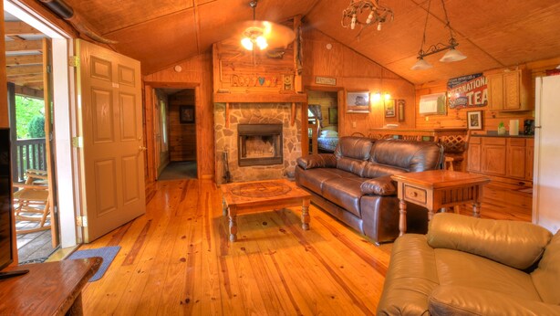 A cozy cabin interior with a stone fireplace, leather sofas, wooden floors, and a kitchen area. - Panther Creek Cabins (Cherokee)