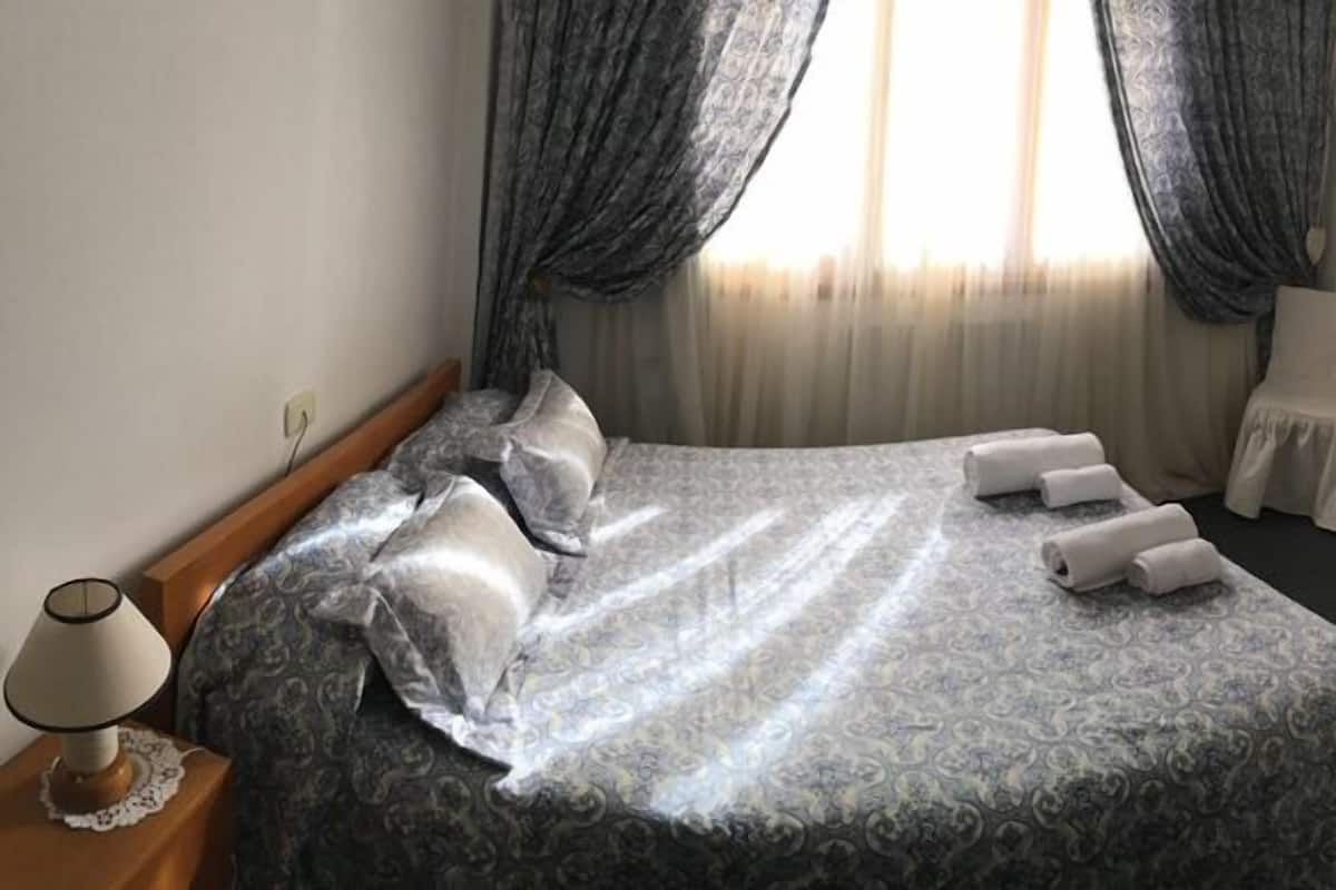 Un lit bien fait, recouvert d’une couvre-lit à motifs, une table de chevet avec une lampe, une chaise, un petit bureau et un miroir accroché au mur.