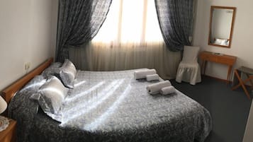 A neatly made bed with a patterned bedspread, a bedside table with a lamp, a chair, a small desk, and a mirror on the wall.