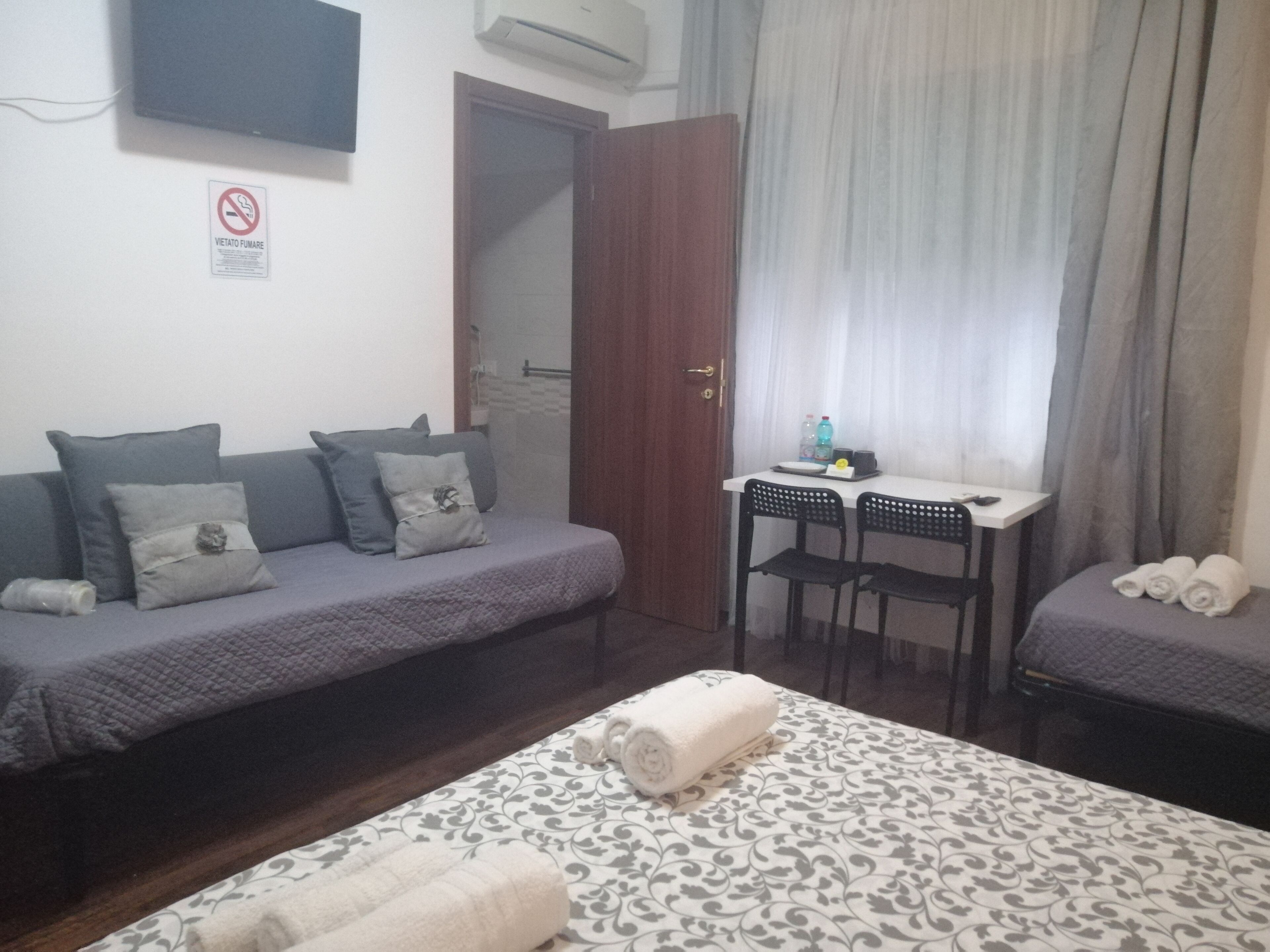 A small, neatly arranged room with a sofa, a bed, a desk with chairs, and a bathroom door.