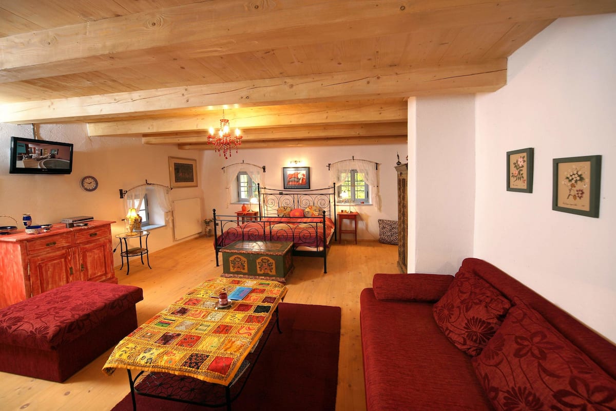 A cozy room with a wooden ceiling, a bed with a canopy, a red sofa, a table with a quilt, and a television on the wall.