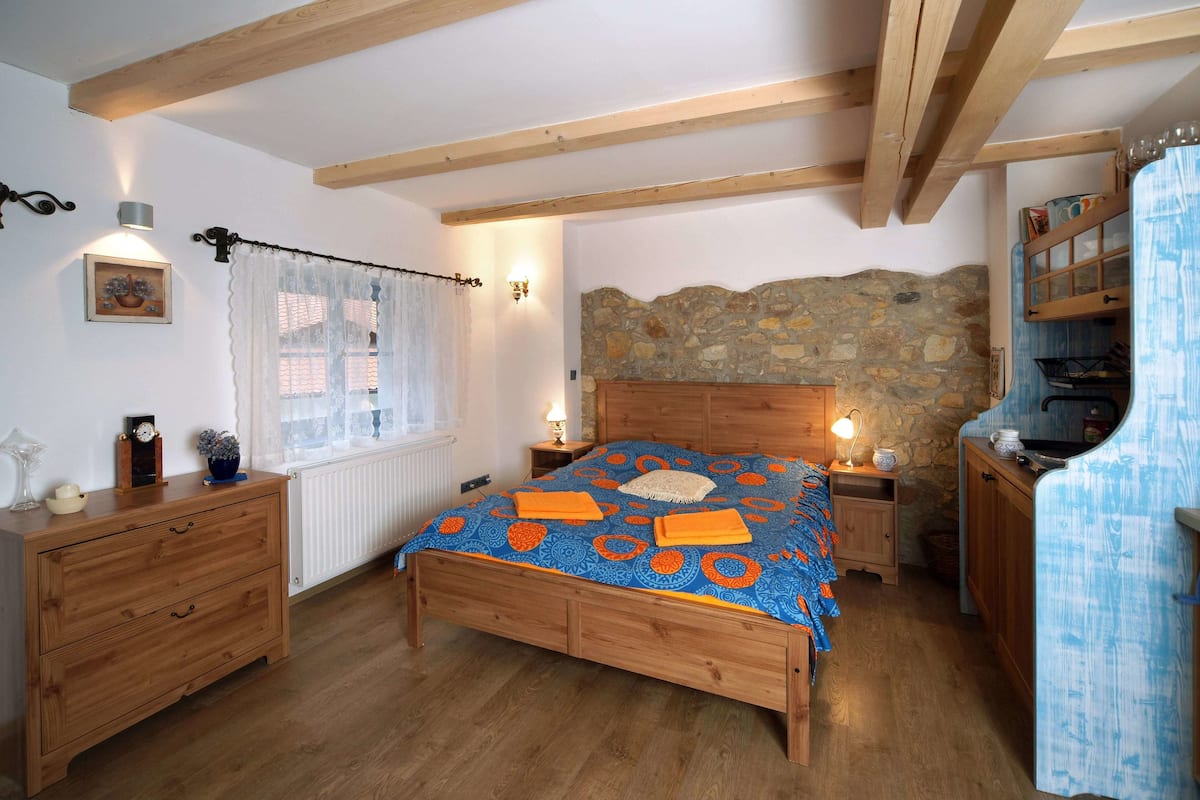 A bedroom with a wooden bed, a stone wall, a window with curtains, a wooden dresser, and a nightstand with a lamp.