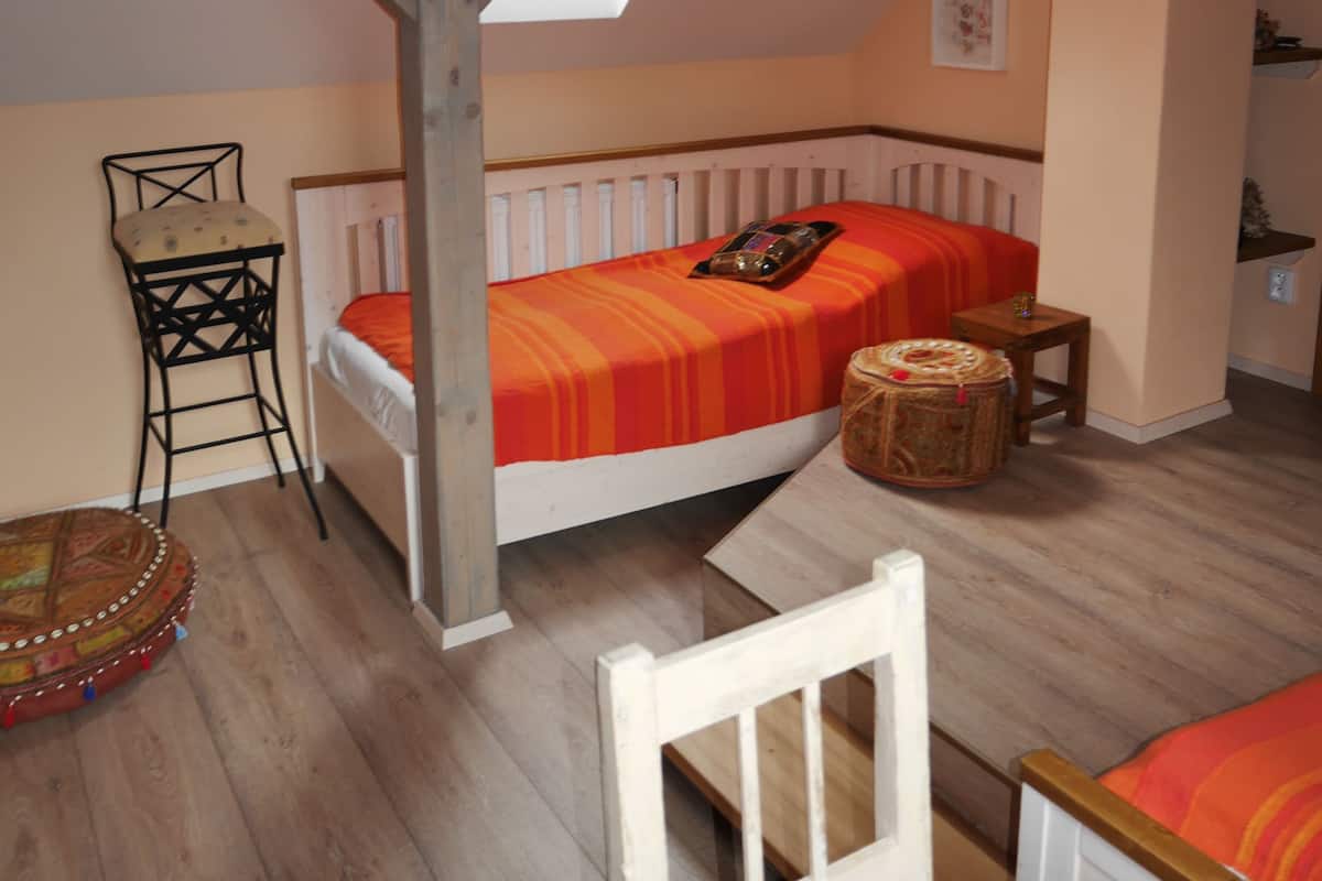 A cozy attic bedroom with a sloped ceiling, a bed with an orange bedspread, a small wooden table, a chair, and a decorative hanging light.